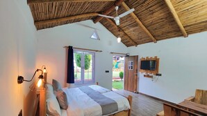 A bedroom with a wooden bed, a desk, a TV mounted on the wall, and a ceiling fan. - The Farm Cottages (Nainital)