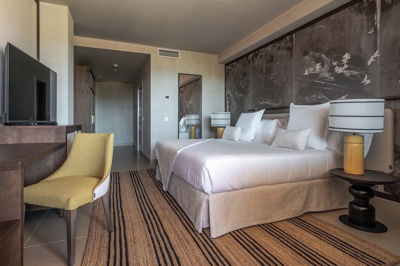 A hotel room with a bed, a chair, a desk, a TV, and a lamp.