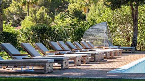 Zona de piscina con sillones y tumbonas dispuestas en filas, una terraza de madera y una piscina.