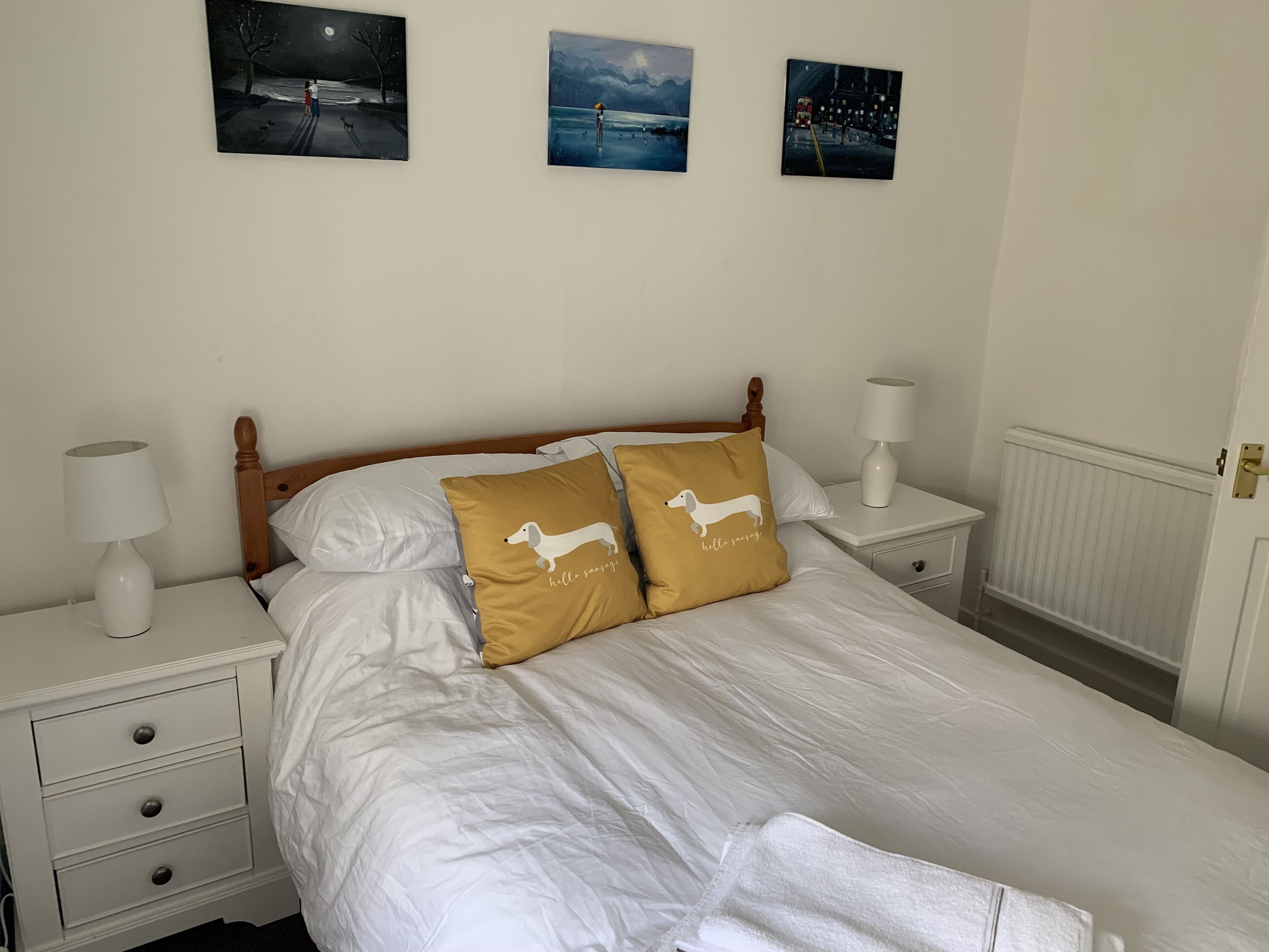 A neatly made bed with white bedding and two pillows, one with a dog illustration and text, in a room with white walls and three framed pictures above the bed.