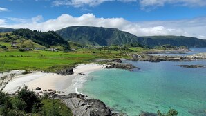 Paesaggio costiero con spiaggia sabbiosa, acque cristalline turchesi e colline e montagne verdi sullo sfondo.