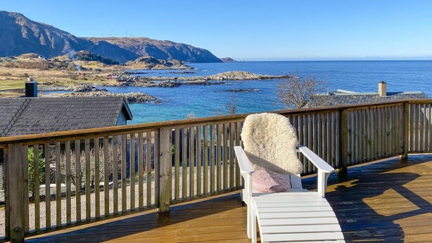 Un terrazza in legno con una sedia a sdraio bianca e un cuscino soffice, affacciata su un paesaggio costiero con scogliere rocciose e pale eoliche all'orizzonte.