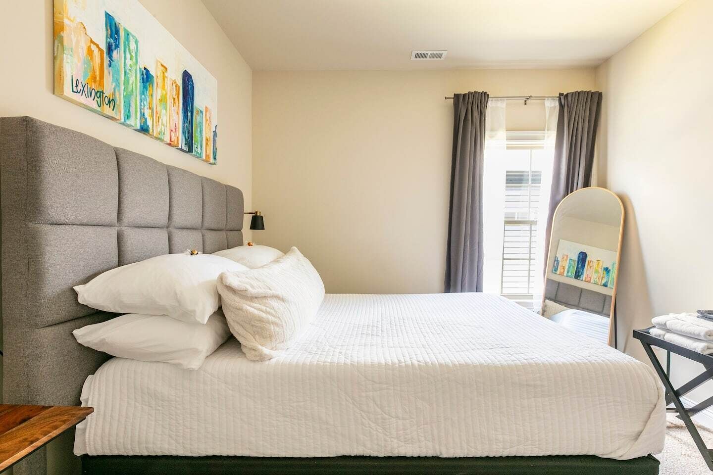 A neatly made bed with white bedding and pillows, a grey upholstered headboard, and a wall-mounted painting of city buildings.