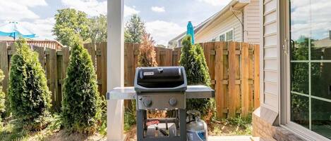 A barbecue grill with a propane tank in a backyard with a wooden fence and trees.