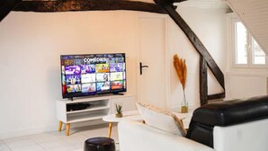 Ein Wohnzimmer mit Fernseher, der einen Comedy-Sender zeigt, einem weißen Sofa, einem schwarzen Ledersessel und einem kleinen Tisch mit einer Pflanze.