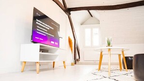 Ein modernes Wohnzimmer mit einem Flachbildfernseher auf einem weißen Standfuß, einem runden Holztisch und einem schwarzen Sessel.
