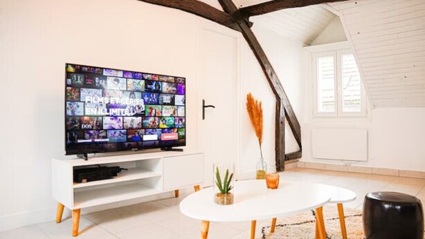 Ein modernes Wohnzimmer mit einem großen Flachbildfernseher an einer weißen Wand, einem weißen Esstisch mit einer kleinen Pflanze und einem schwarzen Hocker.