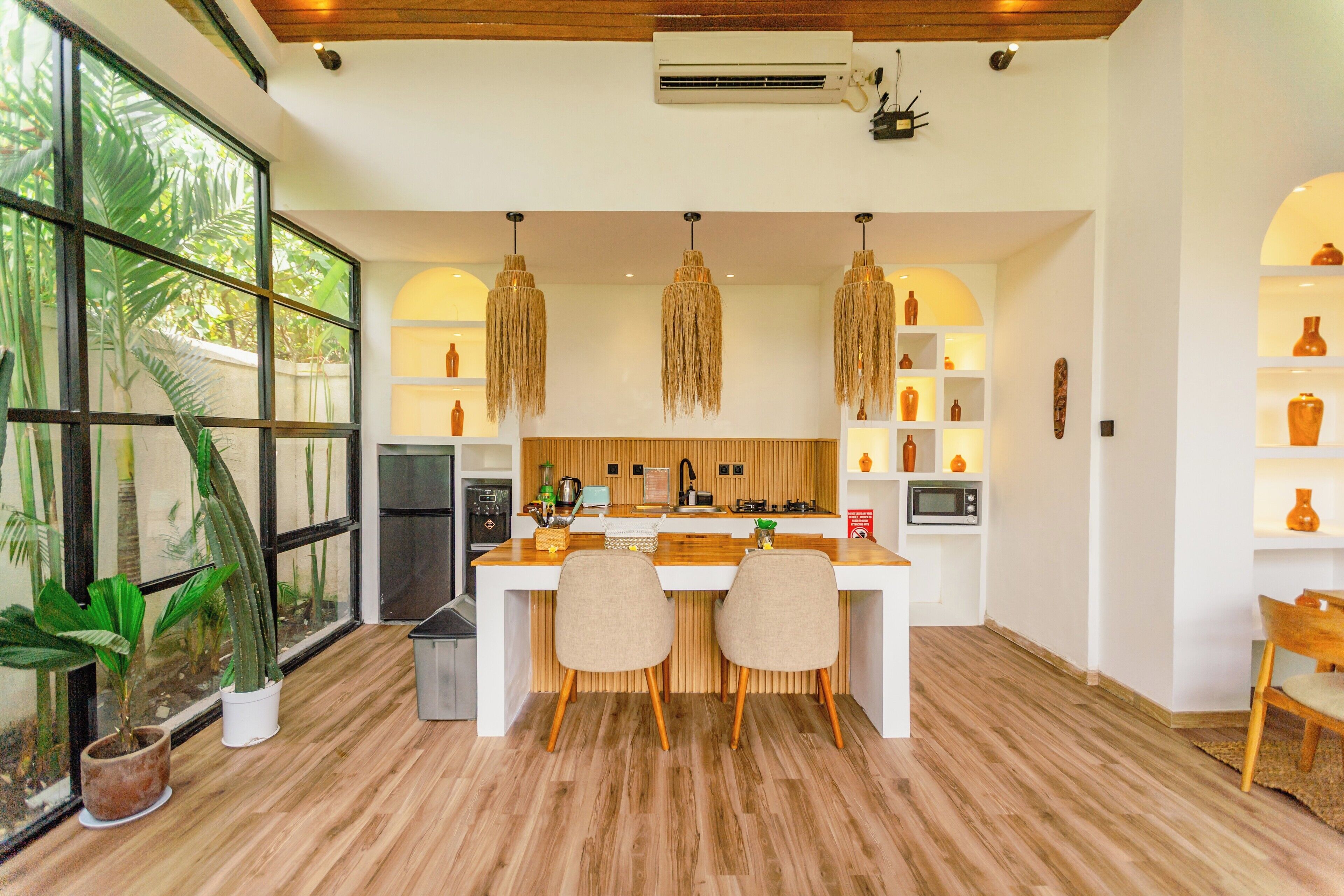 A modern kitchen with a central island, two chairs, and a dining table. There are hanging pendant lights, a microwave, and a stove.