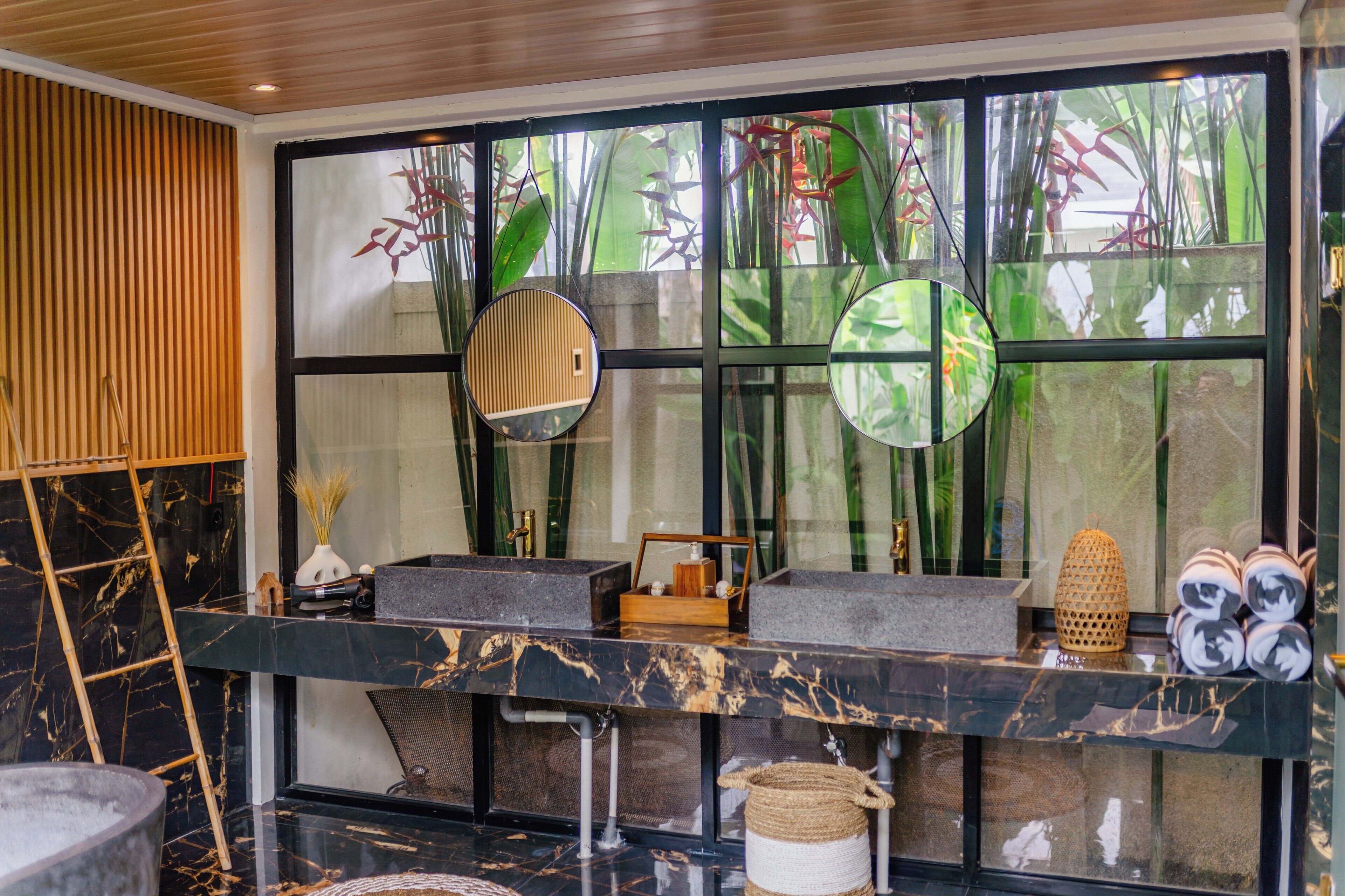 A modern bathroom with a black marble sink, a wooden ladder, and a large window with a view of green plants.