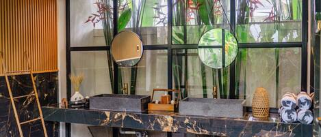A modern bathroom with a black marble sink, a wooden ladder, and a large window with a view of green plants.