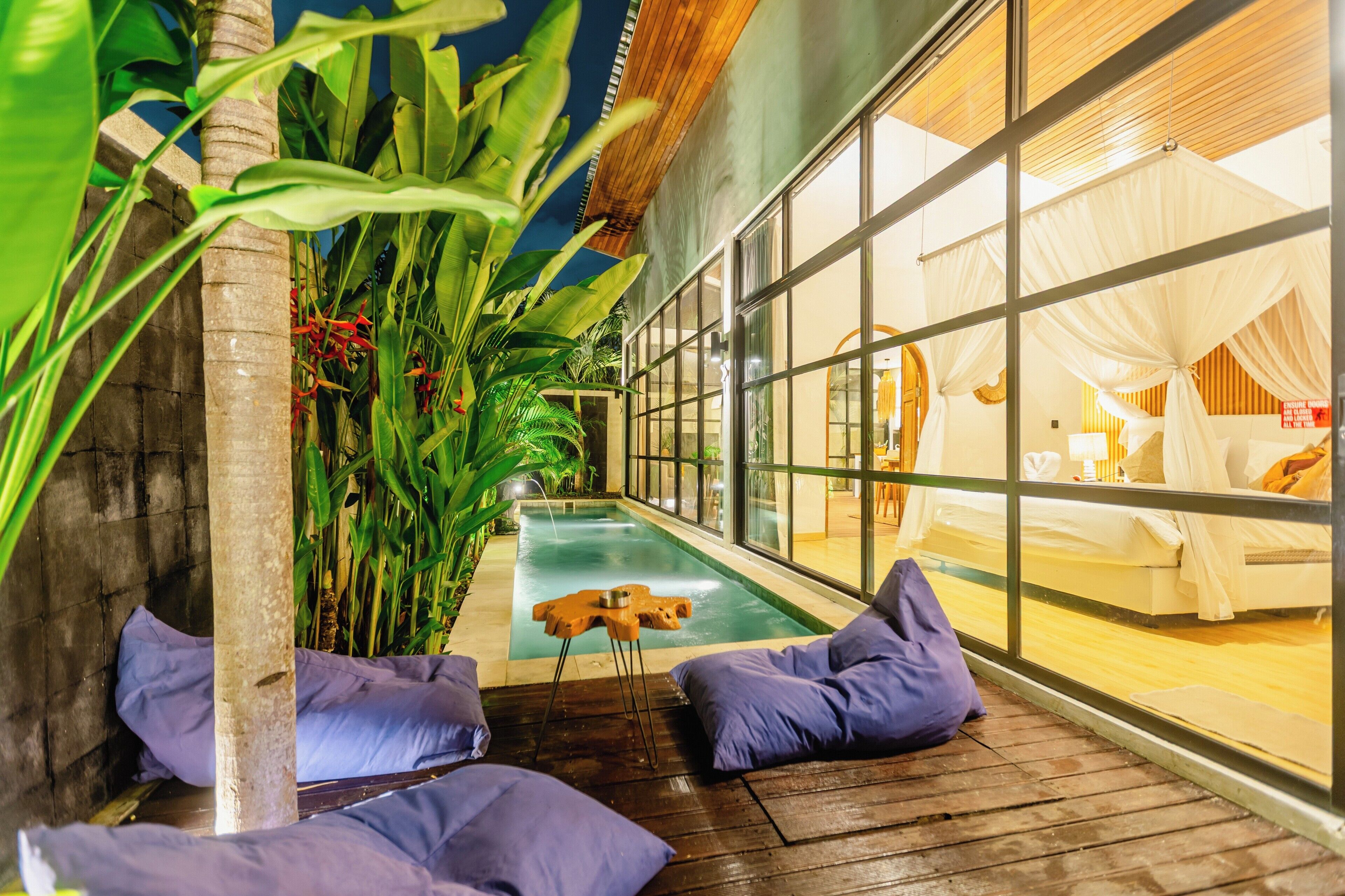 Un espace piscine doté d’une terrasse en bois, de chaises longues et d’une pièce fermée par des parois de verre, comprenant un lit et des rideaux.