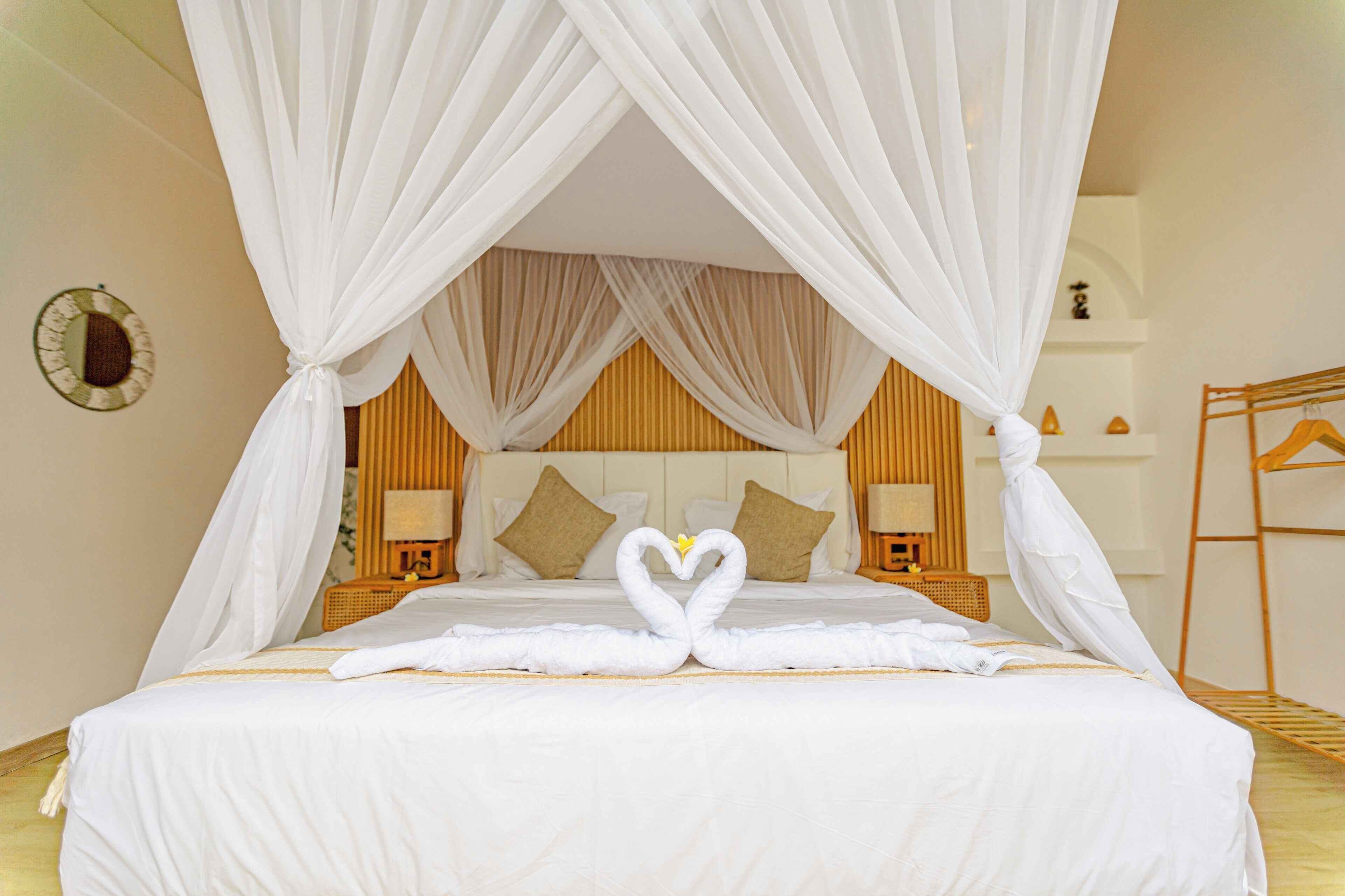 A neatly made bed with a heart-shaped towel arrangement, a wooden headboard, and a bedside table with a lamp.