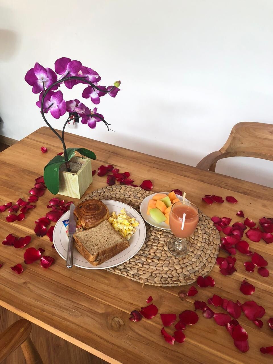 A wooden table with a plate of toast, eggs, and fruit, a glass of juice, and a potted orchid.