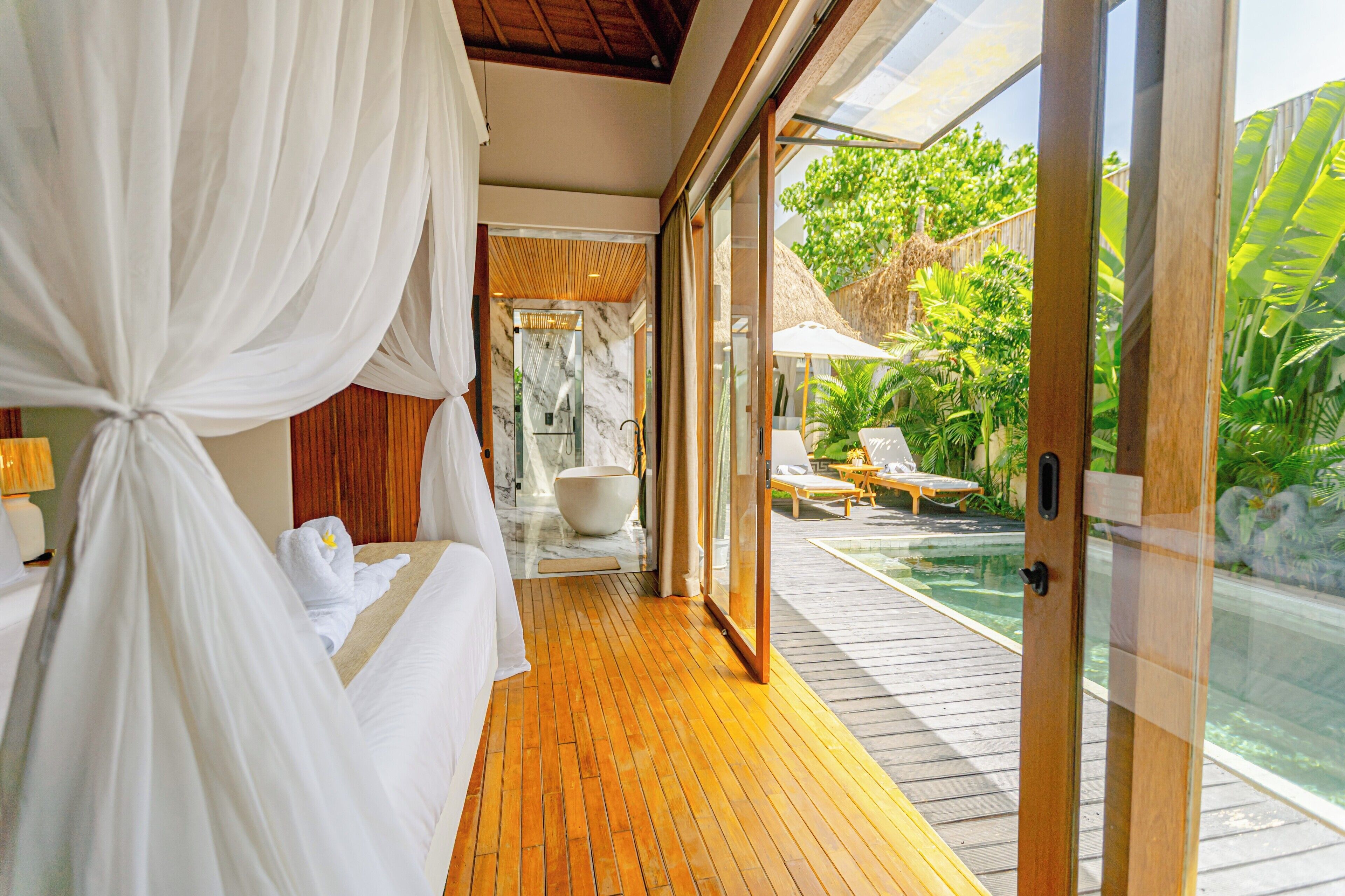 Quarto com piso de madeira, uma cama grande com lençóis brancos e uma porta de vidro que dá para uma área de piscina com espreguiçadeiras.