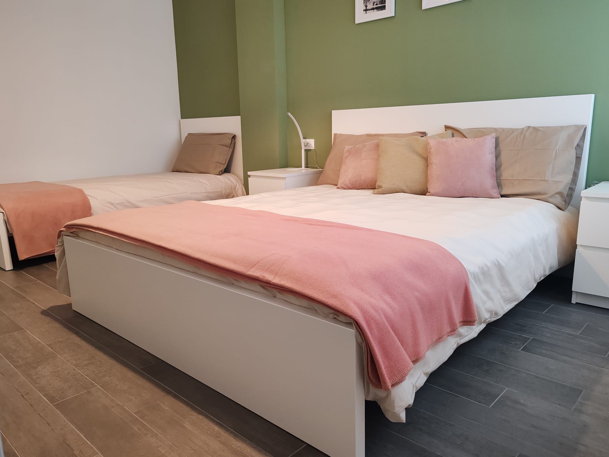 A double bed with a white headboard and a pink blanket, a single bed with a white headboard and a brown pillow, and a nightstand with a lamp.