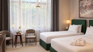 Une chambre d’hôtel avec deux lits, un bureau, une chaise, une petite table ornée d’une fleur et un tableau accroché au mur.