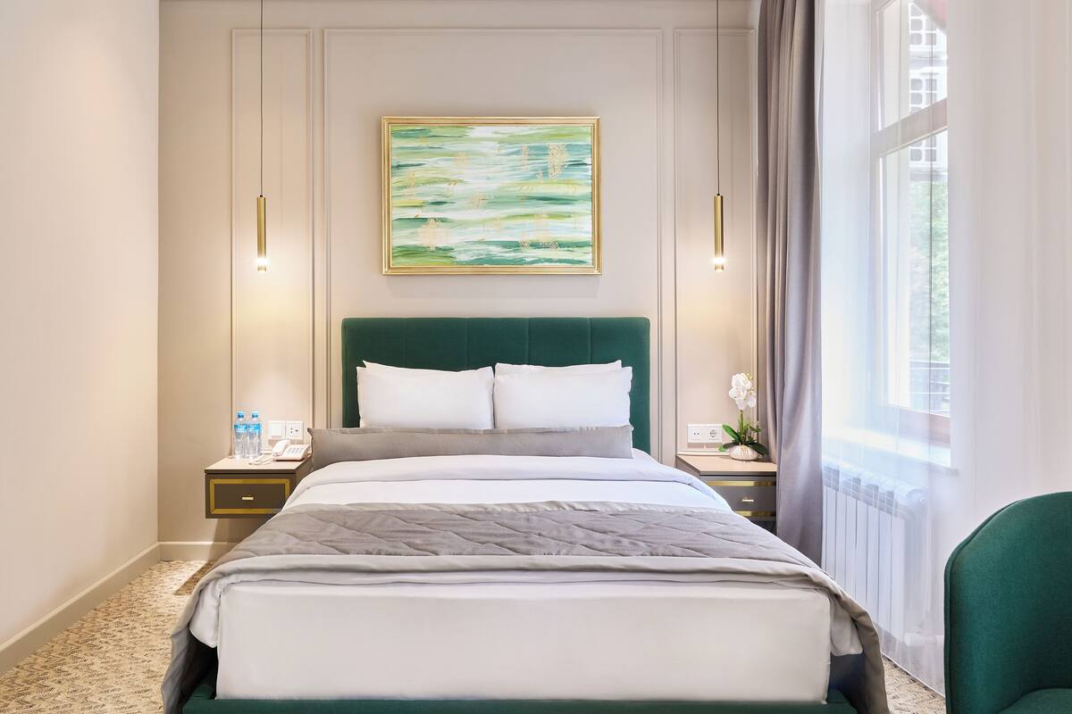 A hotel room with a large bed, a green headboard, a painting on the wall, and a window with curtains.