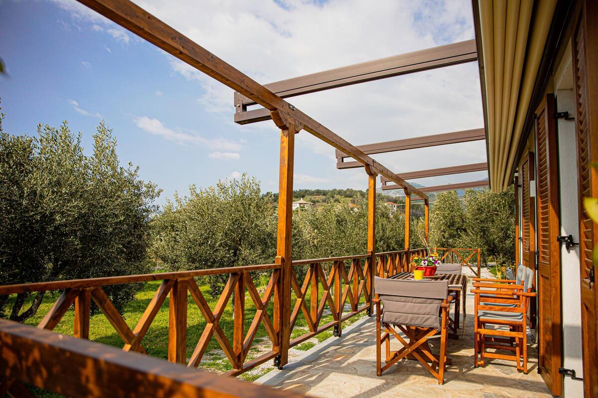 A wooden balcony with a table and chairs, overlooking a garden with olive trees.