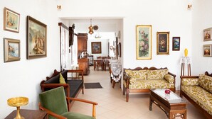 A living room with a green armchair, a patterned sofa, and a wooden coffee table. There are framed artworks on the walls and a chandelier hanging from the ceiling.