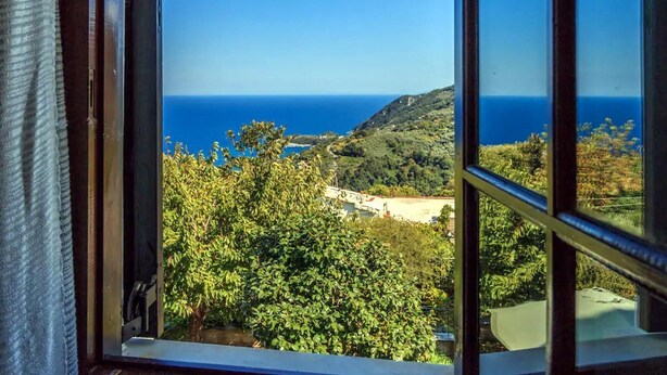 A window view of a coastal landscape with a clear sky, a hill, and a building in the distance. - Apartment Zina Pelion (Zagora-Mouresi)