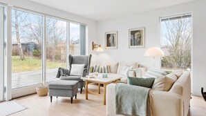 Ein geräumiges Wohnzimmer mit großem Fenster, einem grauen Sessel, einem Holztisch und einer Couch mit gestreiften Kissen.