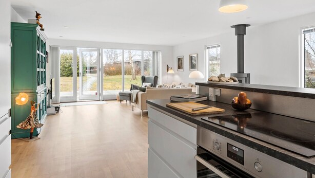Eine moderne Küche mit einer großen Kochinsel, einem eingebauten Backofen und einem Herd. Ein Essbereich mit Tisch und Stühlen lädt zum Verweilen ein, und große Fenster bieten einen Blick ins Freie.
