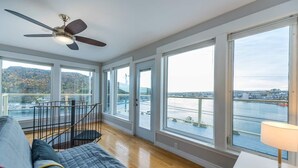 Une pièce spacieuse avec de grandes fenêtres, un ventilateur de plafond, un escalier et une vue sur une étendue d’eau et le paysage urbain.