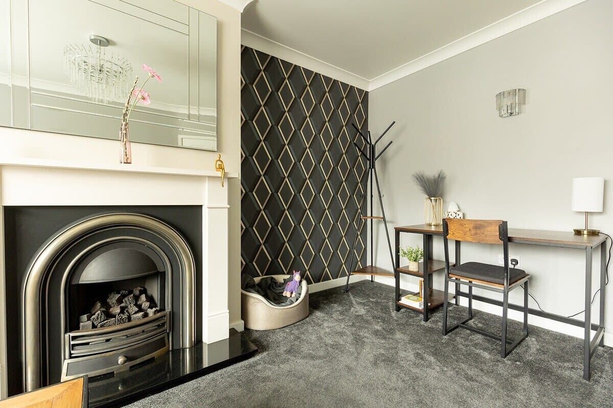 A modern living room with a fireplace, a black diamond-patterned wall, a wooden desk, and a small seating area.
