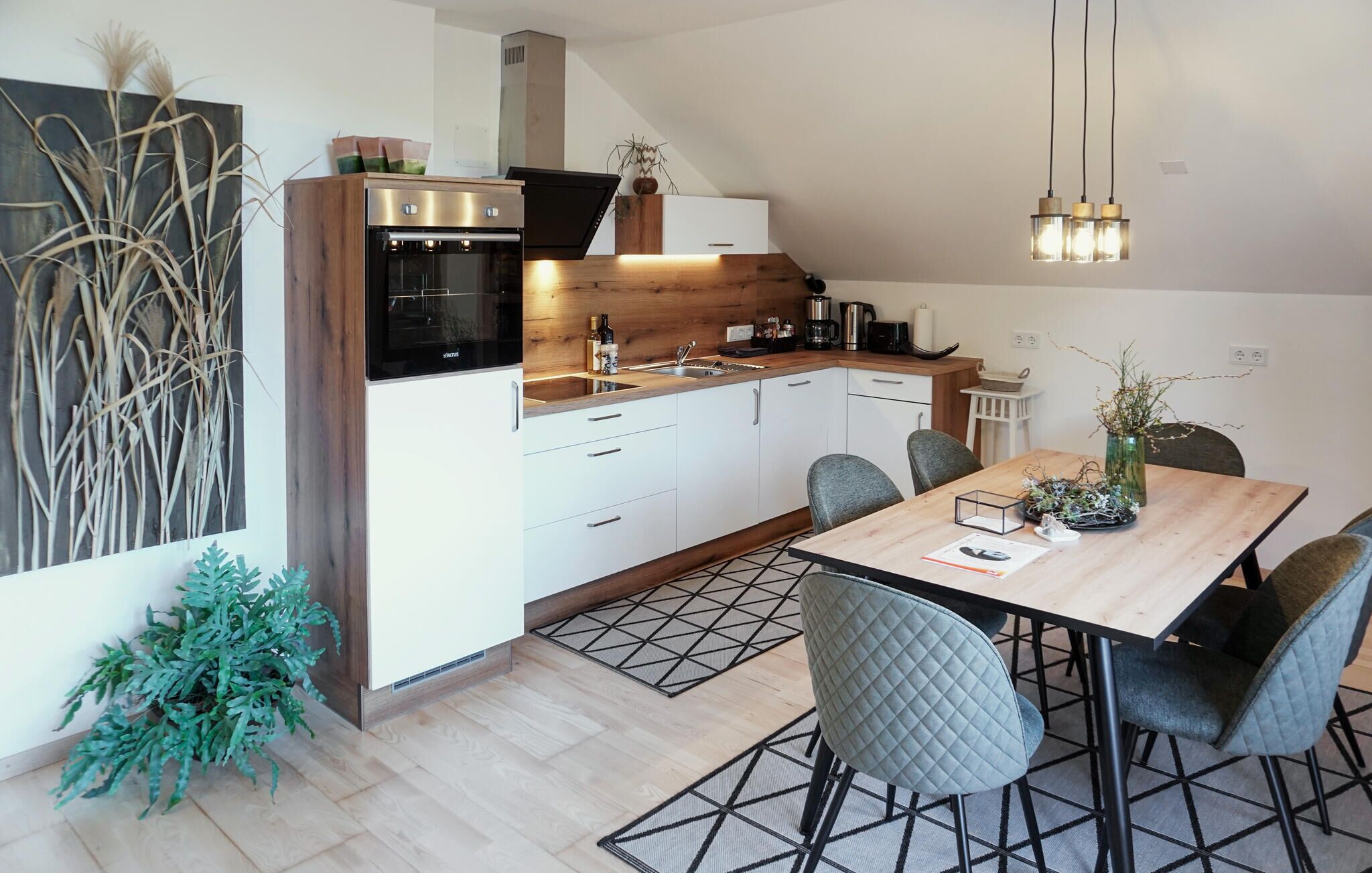 Een moderne keuken met witte kastjes, een ingebouwde oven en een koffiezetapparaat. Er is een eettafel met stoelen en een vaas met takken.