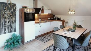 Een moderne keuken met witte kastjes, een ingebouwde oven en een koffiezetapparaat. Er is een eettafel met stoelen en een vaas met takken.
