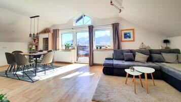 Een ruime woonkamer met een eettafel, stoelen, een bank en een klein bijzettafeltje. Er is een groot raam met uitzicht op bergen.