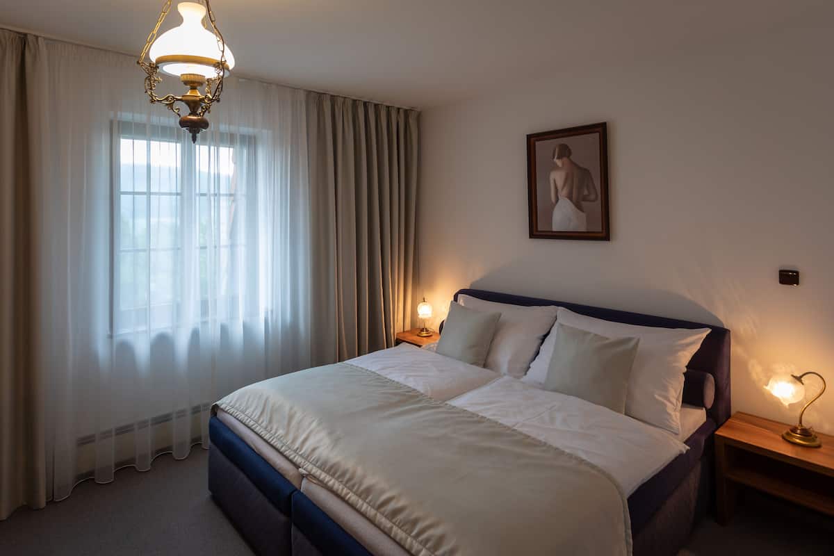 Una cama bien hecha con ropa de cama blanca, dos mesitas de noche con lámparas, un cuadro en la pared y cortinas de encaje en la ventana.