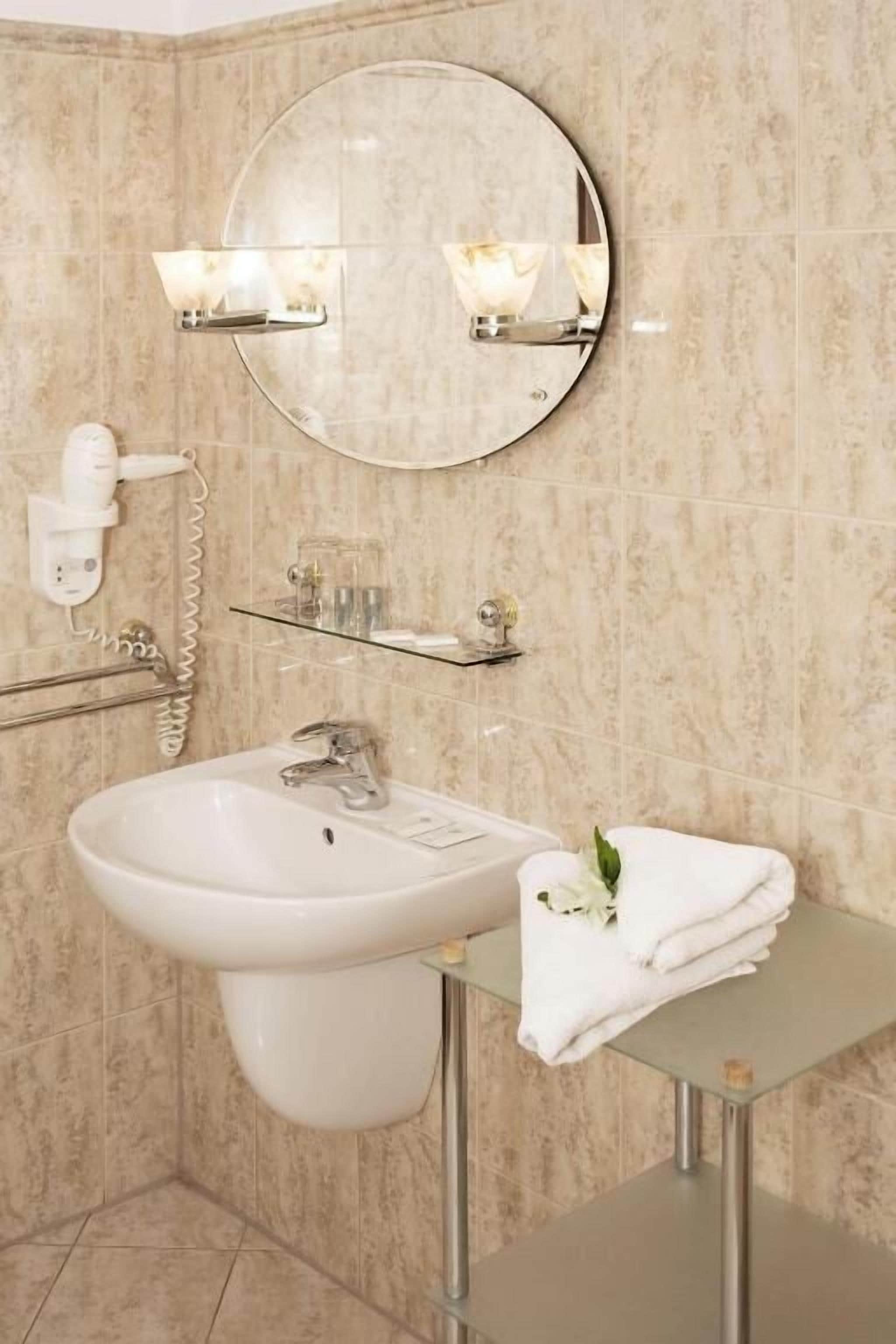 Un baño con un espejo redondo, un lavamanos, un perchero para toallas y un estante de vidrio con artículos de aseo.