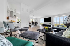 A modern living room with a sofa, ottoman, and coffee table, a dining area with chairs and a table, and a kitchen with a TV and large windows.