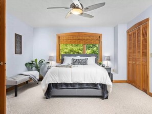 Una camera da letto con un letto, comodini, una panca, una ventola a soffitto, una finestra vista verde e un armadio in legno.