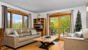 Un salón amplio con un sofá beige, una mesa de centro de madera y una estantería. Grandes ventanales ofrecen vistas al exterior.
