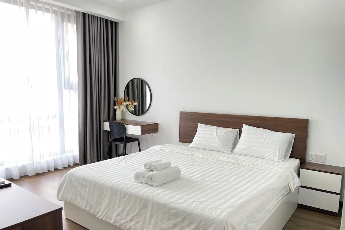 A hotel room with a large bed, a desk with a chair, a round mirror, and a view of the outside through sheer curtains.