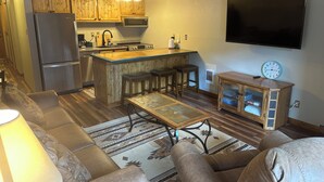 A modern kitchen with wooden cabinets, a stainless steel refrigerator, a microwave, and a large island with bar stools. There is a flat-screen TV mounted on the wall and a clock on the wall.