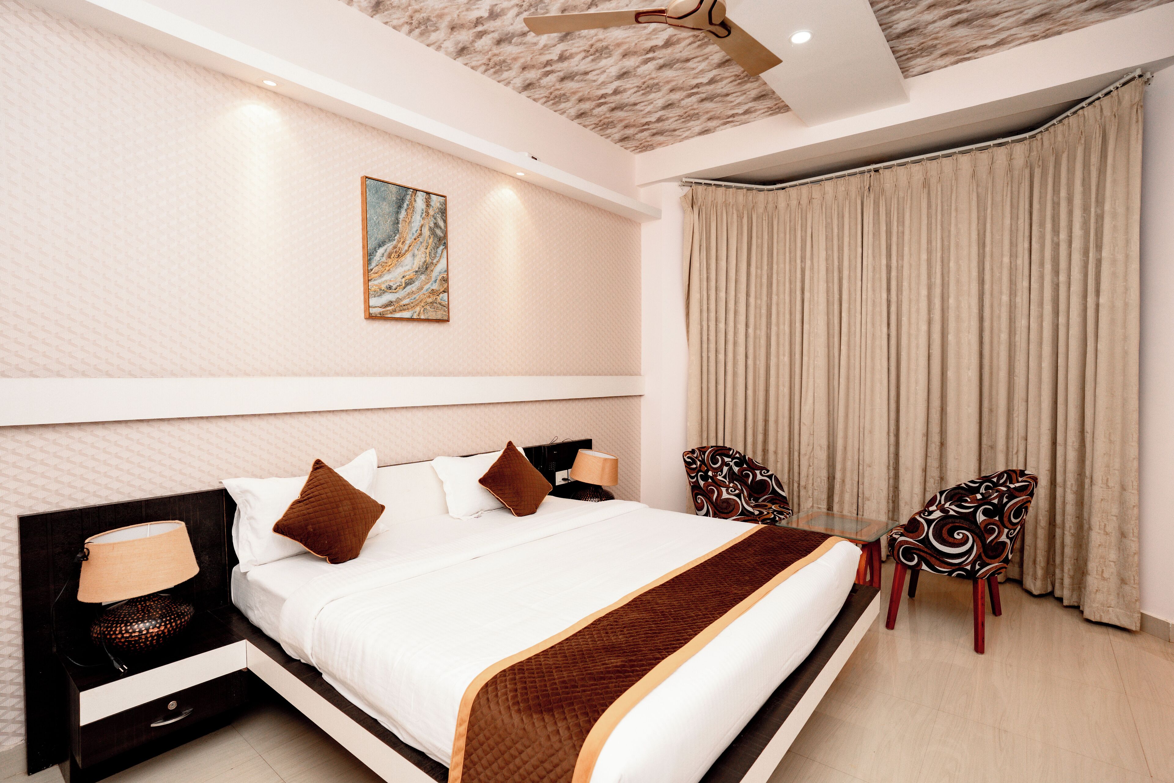 A modern hotel room with a large bed, a nightstand, two chairs, a ceiling fan, and a painting on the wall.