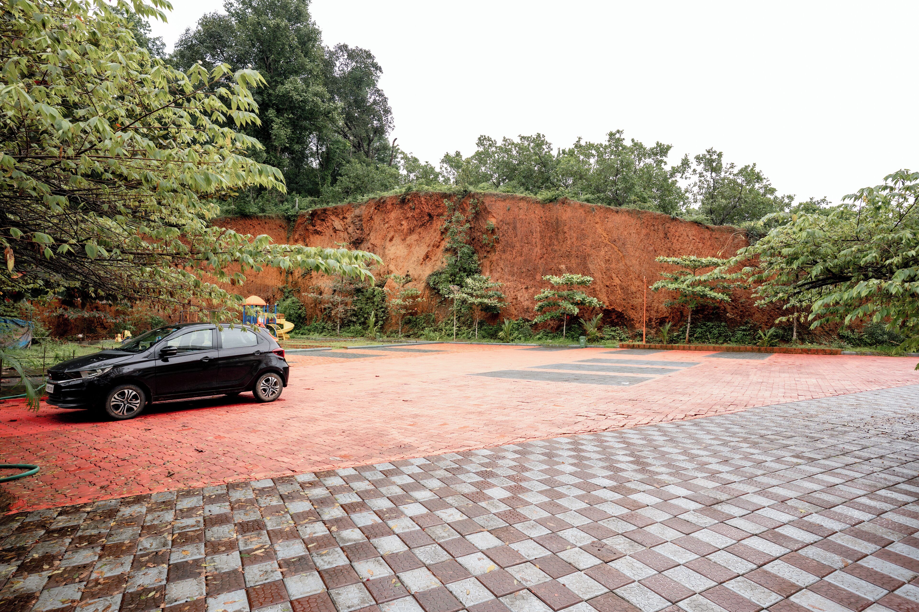 A paved area with a car parked on the left, a red brick path, and a large red hill in the background.
