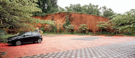 Un área pavimentada con un coche aparcado a la izquierda, un sendero de ladrillo rojo y una gran colina roja al fondo.