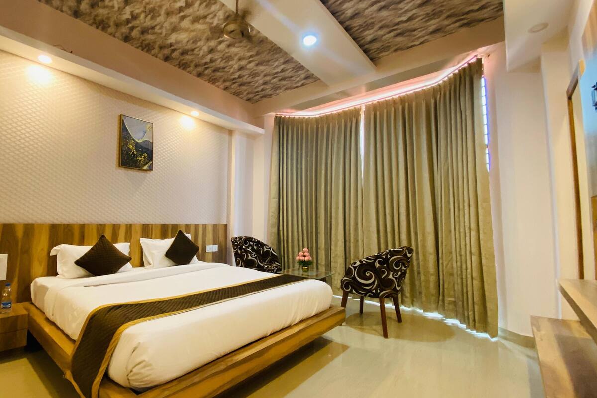 A hotel room with a large bed, a wooden headboard, a nightstand with a lamp, a chair, and a large window with curtains.