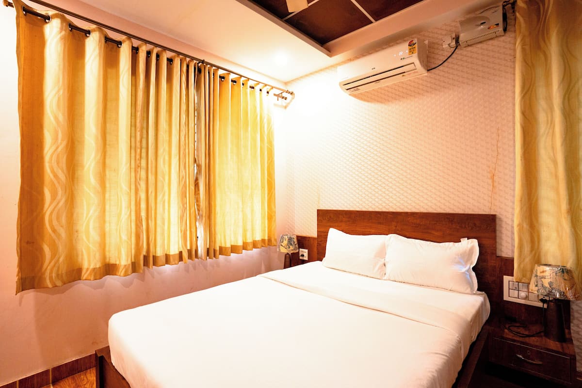 A hotel room with a large bed, a wooden headboard, a nightstand with a lamp, and an air conditioning unit on the wall.