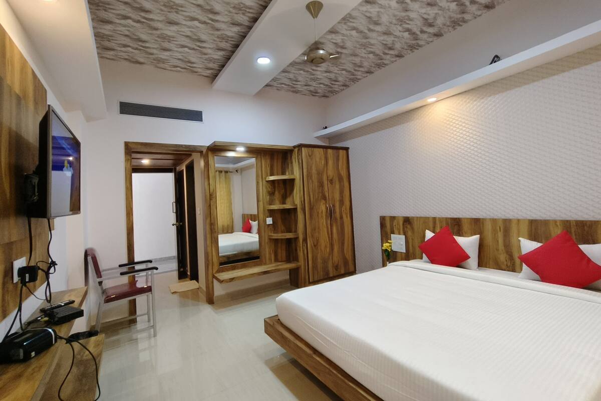 A modern hotel room with a wooden headboard, a large bed with red pillows, a wooden wardrobe, a mirror, and a television mounted on the wall.
