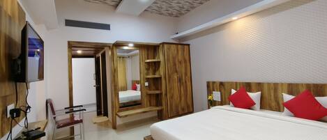 A modern hotel room with a wooden headboard, a large bed with red pillows, a wooden wardrobe, a mirror, and a television mounted on the wall.