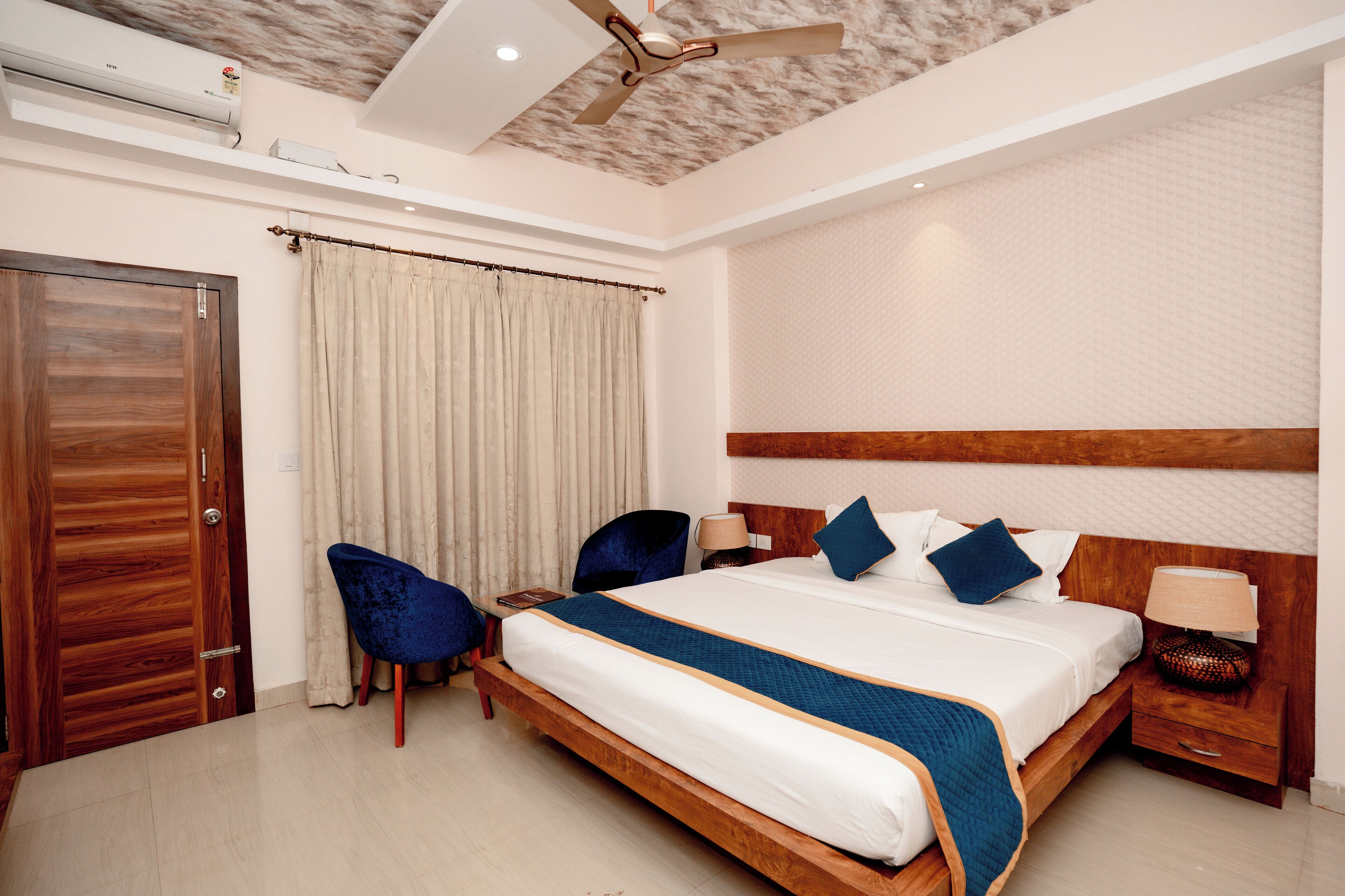 A hotel room with a large bed, a wooden headboard, a ceiling fan, a window with curtains, a wooden door, and a nightstand with a lamp.