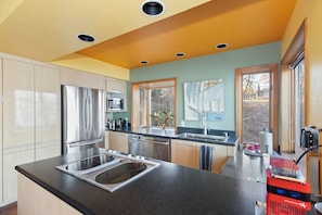 A modern kitchen with stainless steel appliances, a black countertop, and a view of the outside through large windows.