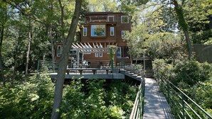 Una casa de madera con varias ventanas y un balcón, rodeada de abundante vegetación y un puente de madera.