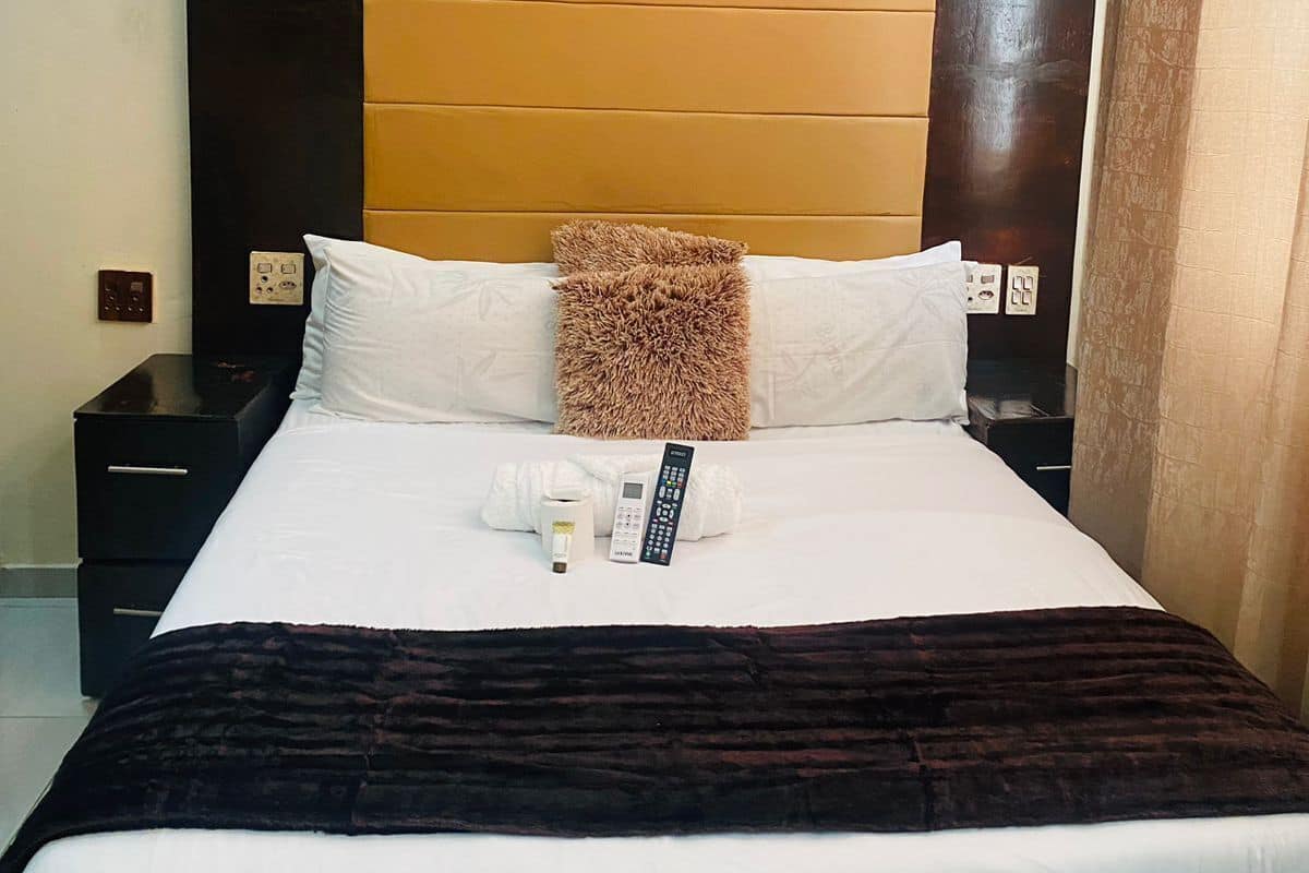 Habitación de hotel con una cama grande, cabecera de madera, ropa de cama blanca, almohada marrón y un frasco de loción, además de un control remoto.