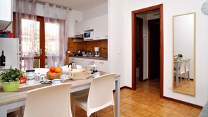 A compact kitchen with white cabinets, a microwave, and a coffee maker. There is a dining table with chairs and a bowl of fruit. - Holiday apartment mit Klimaanlage und Balkon (Lignano)